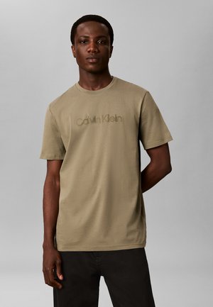 Mężczyzna ubrany w beżowy t-shirt Calvin Klein i czarne spodnie, stojący na tle gładkiego jasnoszarego tła z jedną ręką za plecami.