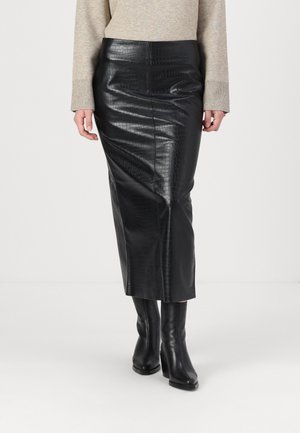 Femme portant une jupe midi noire en cuir synthétique crocodile, un haut beige à manches longues et des bottines à talons noires, debout sur un fond blanc.