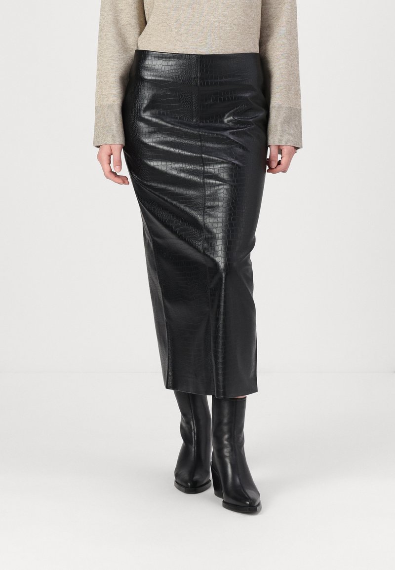 Femme portant une jupe midi noire en cuir synthétique crocodile, un haut beige à manches longues et des bottines à talons noires, debout sur un fond blanc.