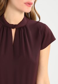 Burgundy blouse with a high neckline, small keyhole opening, and short cap sleeves. Soft, smooth fabric with gathered details.