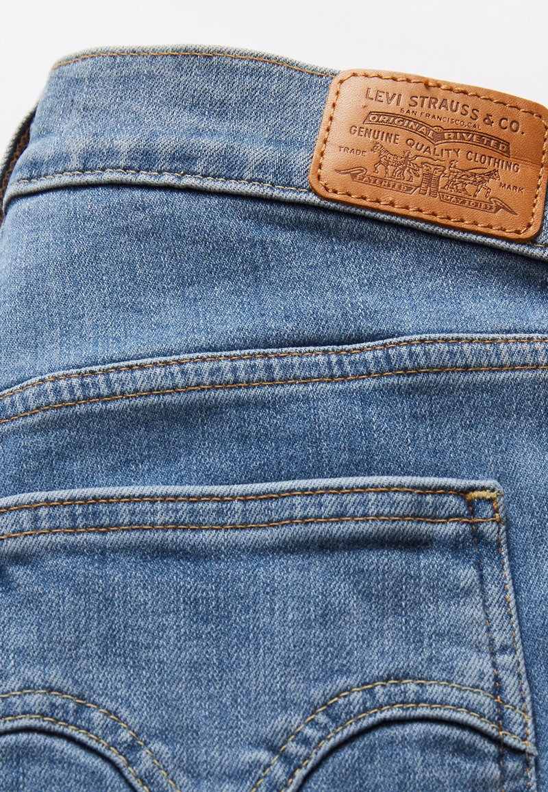 Jean en denim bleu avec une étiquette en cuir arborant un marquage en relief. Couture visible et deux poches arrière avec un détail de design incurvé.