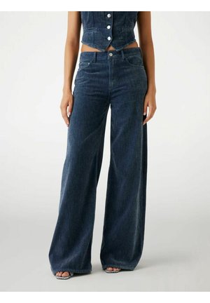 Kvinde iført mørk denimjeans med høj talje og vide ben, kombineret med en ærmeløs denimvest og åbne sandaler.