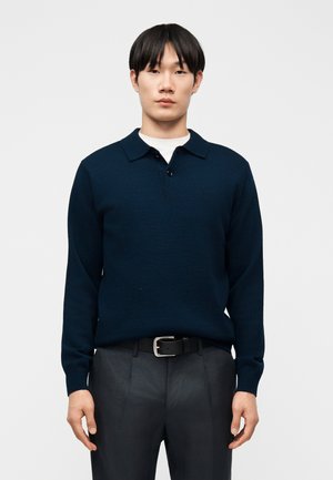 Jeune homme portant un pull à col avec manches longues bleu marine foncé, ceinture noire et pantalon sombre, debout devant un fond gris uni.