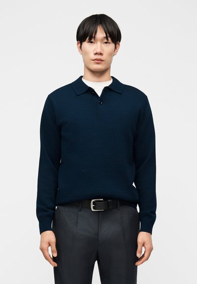 Joven con suéter de manga larga y cuello en tono azul marino oscuro, cinturón negro y pantalones oscuros, posando frente a un fondo gris liso.