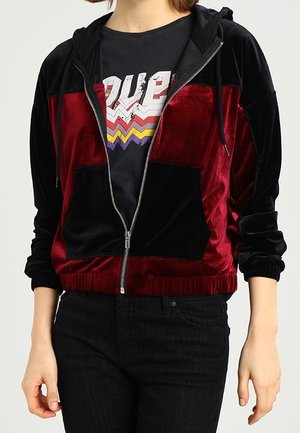 Persona que lleva una sudadera con capucha de terciopelo roja y negra con cremallera sobre una camiseta negra con un texto en zigzag colorido y pantalones negros.