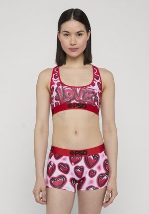 Joven mujer con un conjunto deportivo a juego, sujetador y pantalones cortos con estampado de corazones rojos y rosas y bandas con el logo "PSD", posando frente a un fondo liso.