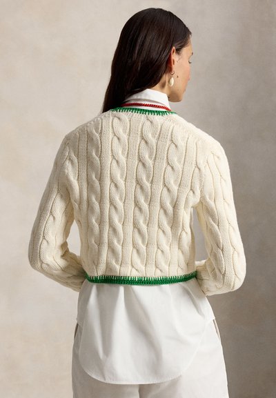 Sieviete ar tumšiem matiem valkā krēmkrāsas īsu džemperi ar pinumu rakstu un zaļu un sarkanu apmali, virs baltas krekla, skatās prom, stāvot pret vienkāršu fonu.