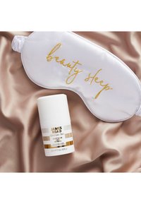 Mascherina da notte in raso bianco con la scritta "beauty sleep" in lettering dorato accanto a un prodotto per la cura della pelle etichettato in bianco e oro su uno sfondo di seta taupe chiaro.
