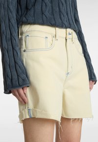 Shorts en denim jaune avec ourlet effiloché, ornés de coutures bleues. Associés à un pull en tricot torsadé bleu foncé.