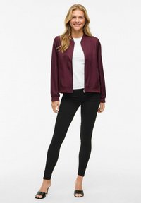 Burgunderfarget sateng bomberjakke med ribbet krage, glidelåslukking og lange ermer, kombinert med en hvit skjorte og svarte leggings.