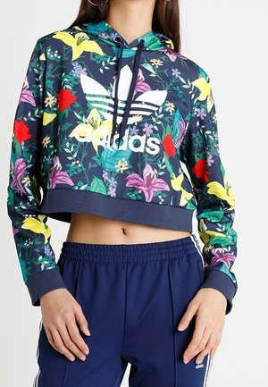 Kobieta nosząca krótką kwiecistą bluzę z kapturem Adidas oraz granatowe spodnie dresowe Adidas, odsłaniająca brzuch i opierająca ręce blisko bioder.