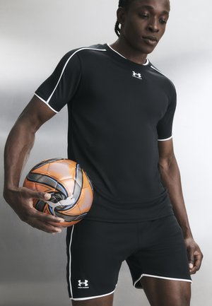 Άνδρας με μαύρα αθλητικά ρούχα Under Armour κρατά μια πορτοκαλί-μαύρη μπάλα ποδοσφαίρου σε απλό γκρίζο φόντο.