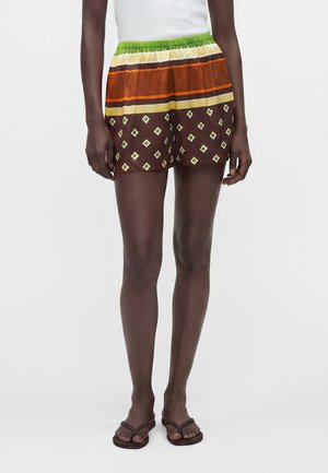 Person iført brune shorts med mønster og grønne, gule og orange striber, en hvid top og mørkebrune flipflops, står på en ensfarvet baggrund.