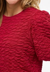 Haut rouge texturé avec un design froissé, encolure ronde et manches bouffantes. Le tissu semble doux et légèrement transparent.