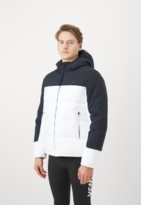 Pufjakke med tofarvet design, hvor den øverste del er marineblå og den nederste del er hvid. Den har lynlås foran, hætte og sidelommer.
