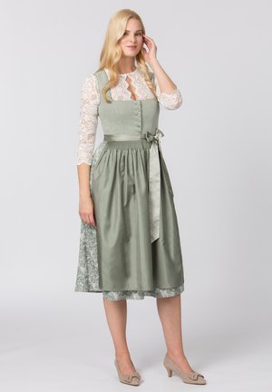 Dirndl-Kleid in gedämpftem Grün mit Spitzenärmeln, Knopfdetails, satiniertem Bogen und gemustertem Rocksaum. Ergänzt durch beige Absätze.