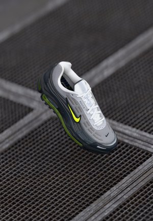 Hellgrauer Sportschuh mit strukturiertem Obermaterial, schwarzen und leuchtend grünen Akzenten, weißen Schnürsenkeln und einer schwarzen Laufsohle mit Dämpfungseigenschaften.