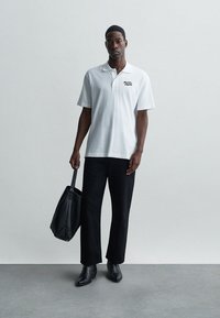 Λευκό πουκάμισο πόλο με κοντά μανίκια, μαύρο κεντημένο κείμενο στο στήθος, συνδυασμένο με μαύρα παντελόνια και μυτερά μαύρα παπούτσια, κρατώντας μια μαύρη τσάντα tote.