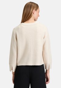 Kremowy, fakturowany top o luźnym kroju, z zagiętym kołnierzykiem i bocznymi rozporkami przy rękawach, zestawiony z czarnymi spodniami.