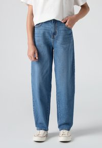 Oseba v svetlo modrih ravnih hlačah z eno roko v žepu in belimi športnimi copati na enobarvni svetli ozadju.