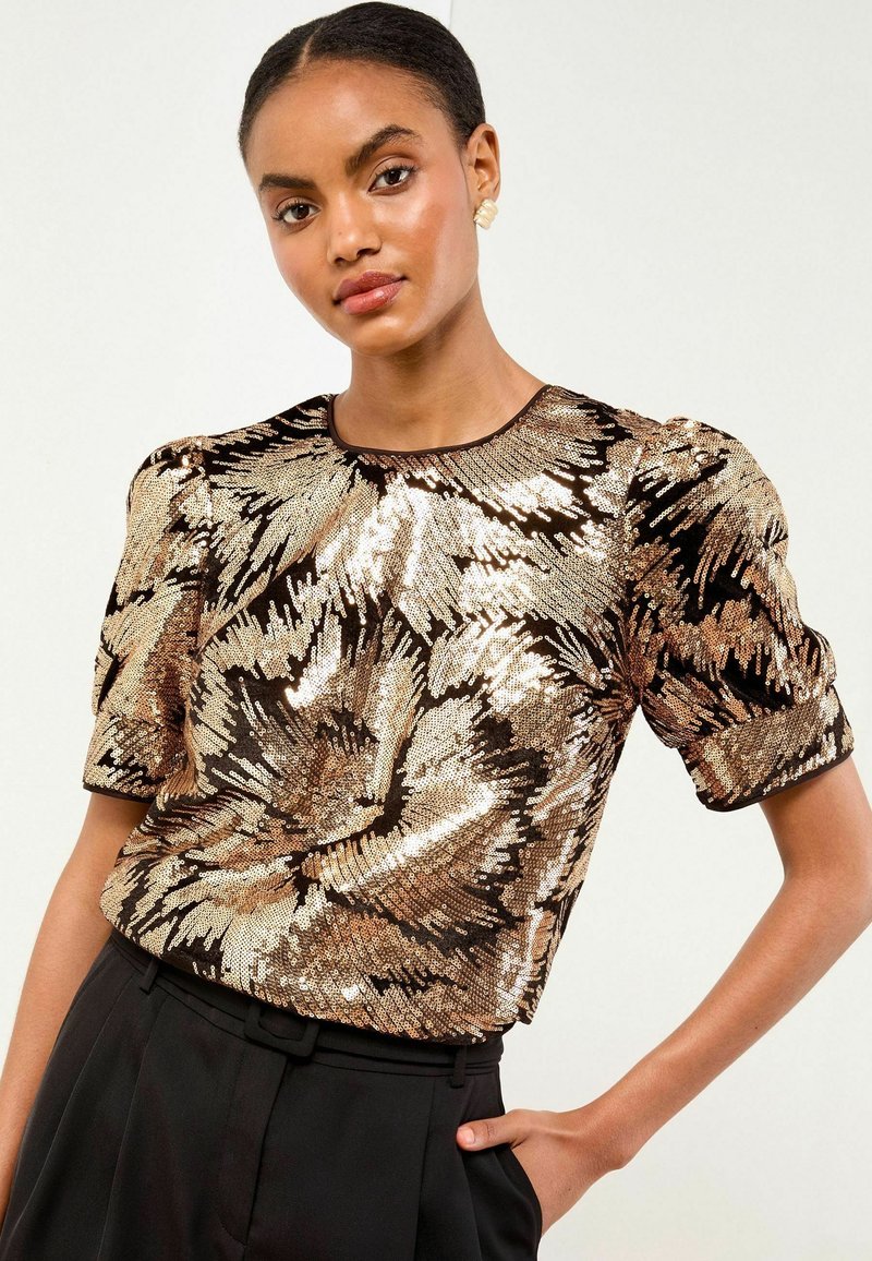 Top a maniche corte in paillettes nere e dorate, con un motivo floreale, scollatura rotonda e spalle plissettate. Indossato con pantaloni neri.