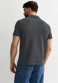 Hombre de cabello corto con una camisa polo de punto de manga corta de color gris oscuro y vaqueros azules, visto desde atrás frente a una pared lisa.
