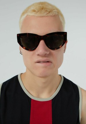 Occhiali da sole cat-eye in tartaruga con lenti scure, montatura spessa e dettagli metallici sulle tempie, indossati da una persona con capelli corti biondo platino.