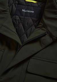 Gros plan d'une veste matelassée vert olive foncé avec fermeture éclair, boutons-pression, poche à rabat et étiquette "Matinique" à l'intérieur du col.