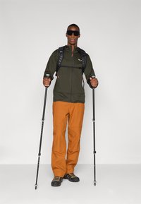 Udendørsudstyr: mørkegrøn zip-up jakke, orange bukser, sorte støvler, rygsæk og vandrestave. Solbriller fuldender looket.