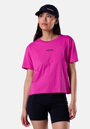 Kvinde iført en skrigende pink Rossignol t-shirt, sorte cykelshorts og en sort Rossignol kasket, kigger let til siden.