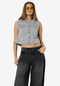 Sivi skraćeni traper prsluk bez rukava s dugmadi, uparen s labavim crnim trapericama. Model nosi sunčane naočale s žutim staklima.