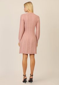 Rosa gestricktes Kleid mit Kabelmuster, knielangem Design, langen Ärmeln und geripptem Kragen. Kombiniert mit schwarzen High-Heels.