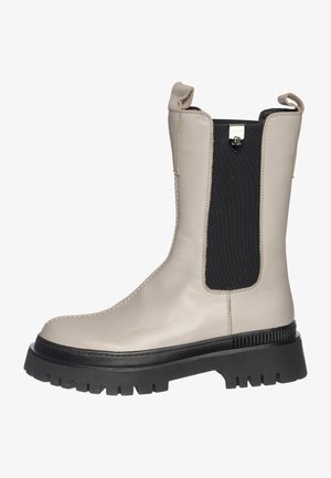 Bota de cuero beige hasta el tobillo con elástico negro en el lateral, suela negra resistente y tira para tirar. Presenta costuras a lo largo de la puntera y acabado liso.