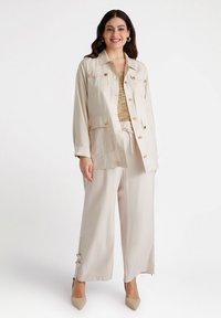 Giacca beige leggera con due tasche sul petto e bottoni dorati; abbinata a pantaloni a gamba larga coordinati e un top neutro dalla texture.