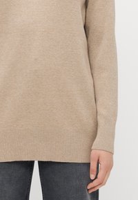 Pull en tricot beige avec un ourlet et des poignets côtelés, présentant une texture douce et une coupe décontractée, associé à un jean en denim noir.