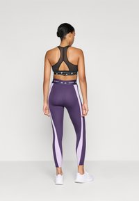 Sujetador deportivo negro con espalda nadadora y panel de malla; leggings morados con rayas laterales en morado claro y logo; zapatillas deportivas blancas.