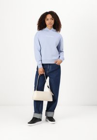 Svetlo modra kapuca s Calvin Klein logotipom, ohlapen jeans, črne superge in majhna bež torbica. Gladek material, sproščen dizajn.