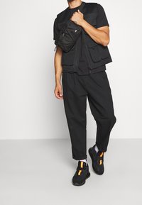 Svart väst med flera fickor, svart t-shirt, lösa svarta byxor och svarta sneakers med orange detaljer. Enkel, funktionell design.