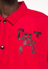 Rode jas met een spits kraag, zwarte knopen en opdrukken waaronder "CHI" en een stierlogo. Glad textuur en sportief ontwerp.