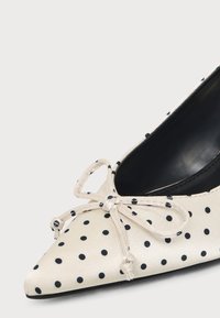 Chaussure plate crème à bout pointu avec des pois noirs et un nœud noué sur le dessus, avec une doublure intérieure noire.