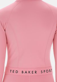 Rózsaszín hosszú ujjú sportfelső, sima textúrával, sötét betűzéssel a derékrészen. Varrott díszítések a kézfejeken és a háton.