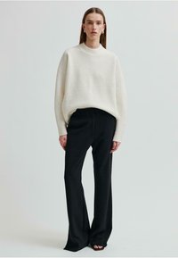 Maglione overdimensionato color crema a maglia con collo alto abbinato a pantaloni neri a vita alta. Tessuto morbido e vestibilità rilassata, con maniche lunghe.