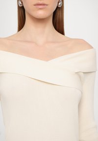 Top de ombro caído em creme, com decote diagonal e mangas longas, feito de um tecido suave e texturizado. Brincos pendentes prateados incluídos.