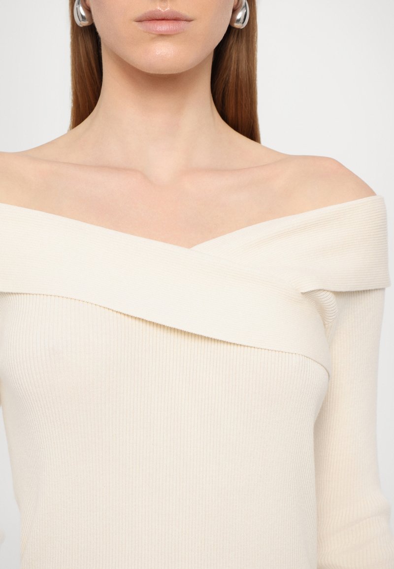 Rippentop mit schulterfreiem Design in Creme, ausgestattet mit einem diagonalen Ausschnitt und langen Ärmeln, aus weichem, strukturiertem Stoff. Inklusive silberner Creolen.