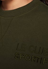 Sudadera verde oliva con cuello redondo, con texto bordado tonal "LE CLUB SPORTIF" y detalles de costura visibles en el cuello.