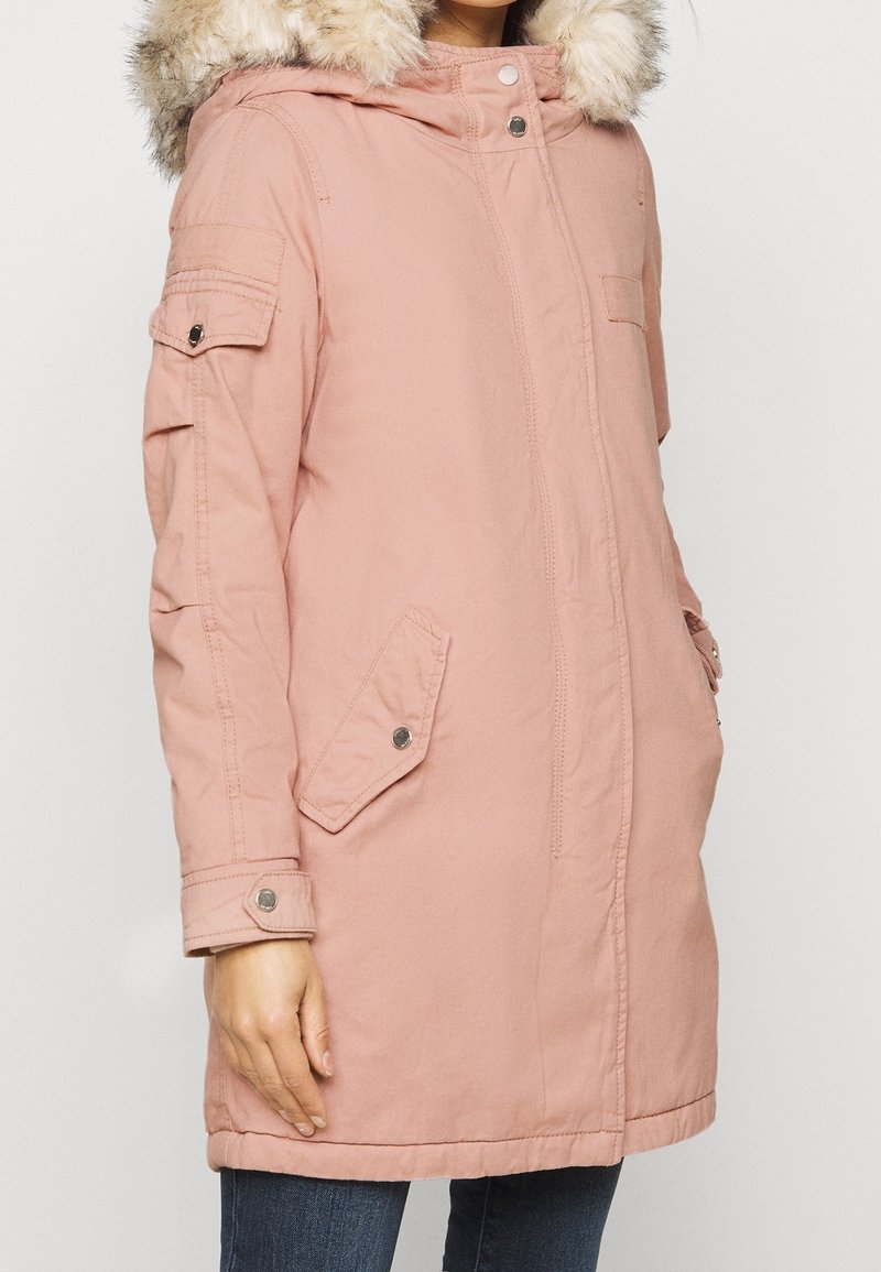 Růžová parka s kožešinovou kapucí, zapínání na zip a knoflíky, boční kapsy a strukturovaná tkanina. Má rukávy s detaily na knoflíky.