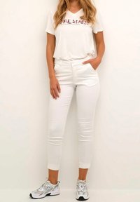 Witte aansluitende broek gecombineerd met een beige T-shirt met het woord "DREAMER" in kleurrijke letters. Schoeisel: grijze en zwarte sportieve schoenen.