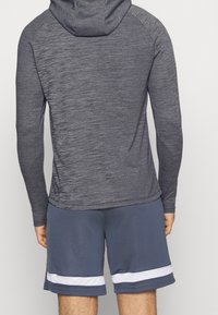 Dunkelgrauer langärmeliger Kapuzenpullover aus strukturiertem Stoff, kombiniert mit blauen Sportshorts, die am Saum einen weißen horizontalen Streifen aufweisen.