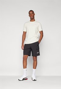 Camiseta color beige claro con un pequeño logo, combinada con pantalones cortos cargo negros que tienen bolsillos laterales. Llevado con zapatillas deportivas blancas y calcetines.