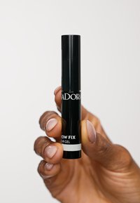 Tubetto cilindrico nero di gel per sopracciglia con etichetta di testo bianca. Il tubetto è tenuto in verticale in una mano con un tono di pelle neutro su uno sfondo bianco.